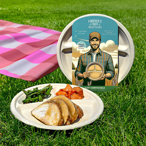 A Better Earth 10 3 Comp Farmers Fiber Collection Round Plate with turkey, green beans, and potatoes sits on grass next to its box featuring a smiling man holding the plate. A pink picnic blanket is visible in the background.