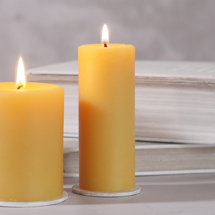 Two 100% Pure Beeswax Pillar Candles by Big Dipper Wax Works glow warmly on a light surface before stacked white books, adding an eco-friendly, toxin-free touch to your home against a soft, neutral background.