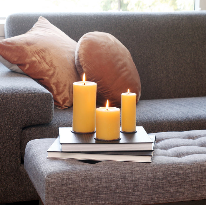 Three 100% Pure Beeswax Pillar Candles by Big Dipper Wax Works of varying heights sit atop stacked books on a gray ottoman, creating a cozy, toxin-free ambiance in front of a gray sofa and window with blurred greenery outside.