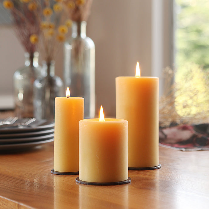 Three lit Big Dipper Wax Works 100% Pure Beeswax Pillar Candles of different heights sit on a wooden table with plates and dried flowers nearby, creating a cozy, toxin-free atmosphere in natural sunlight.