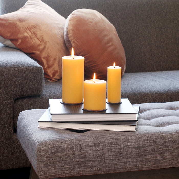 Three 100% Pure Beeswax Pillar Candles by Big Dipper Wax Works glow atop stacked books on a gray ottoman in a modern living room. A gray sofa with round tan pillows completes the cozy, toxin-free ambiance.