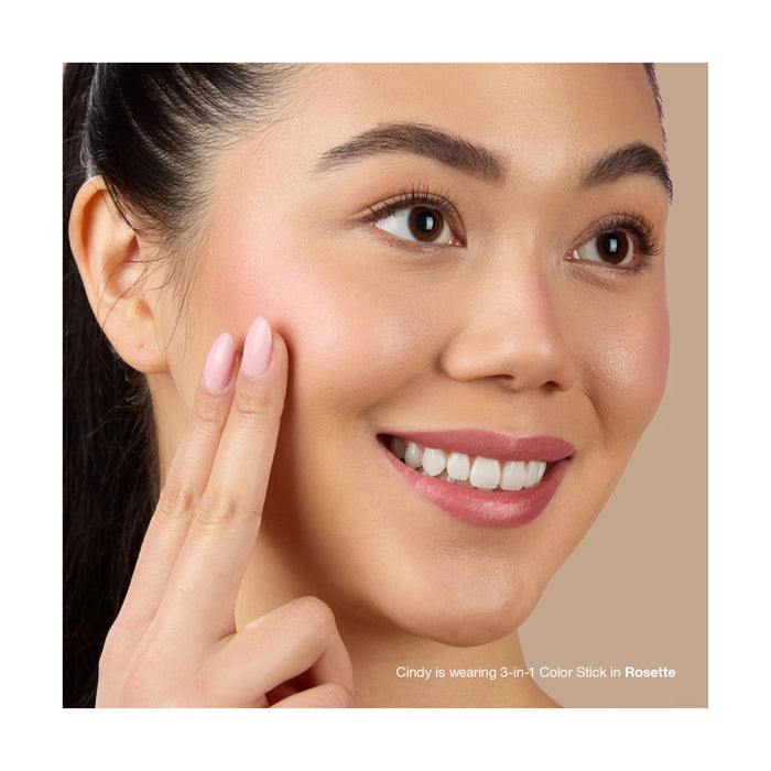 A woman with long dark hair smiles as she applies Mineral Fusion 3-in-1 Color Stick in Rosette to her cheek with her fingers. Her natural makeup has soft pink tones. Text at the bottom reads, Cindy is wearing 3-in-1 Color Stick in Rosette.