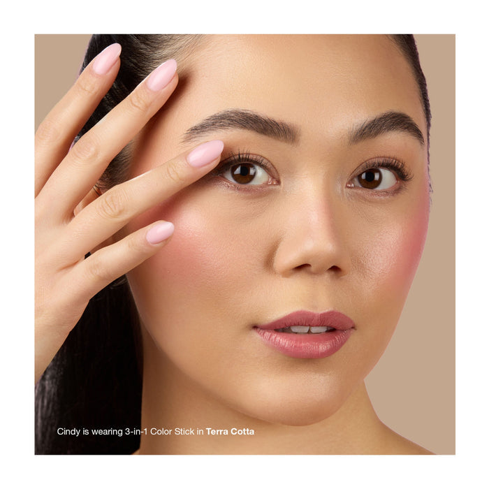 A woman with smooth, radiant skin and pink makeup on her cheeks and lips gently touches her face, highlighting a natural look. Text reads: “She’s wearing Mineral Fusion 3-in-1 Color Stick in Terra Cotta.”.