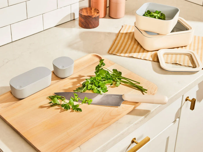 The Caraway 4 Piece Cutting Board Set from Caraway is displayed on a kitchen counter with a large knife and fresh parsley, surrounded by stacked storage containers and a striped cloth near a white tiled backsplash.