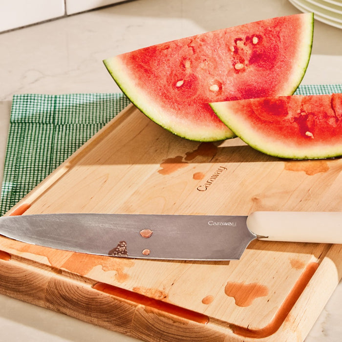 A non-toxic Caraway 4 Piece Cutting Board Set with a large chefs knife and watermelon slices. Watermelon juice is on the knife and board, which rests on a partially visible green placemat.
