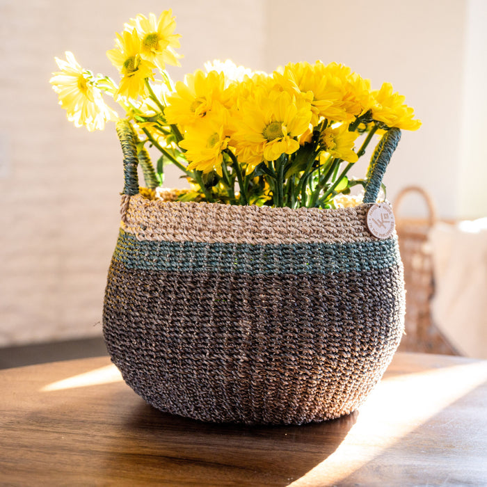 The LIKHA Abaca Bulge Basket, handwoven from ethically sourced Manila hemp with green trim, holds bright yellow flowers and rests on a sunlit wooden surface.