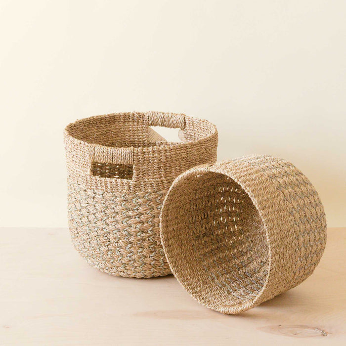 The LIKHA Abaca Round Bottom Basket, a nested set of 2, is shown on a light wooden surface—one upright and one on its side—highlighting their ethically made design and intricate texture.
