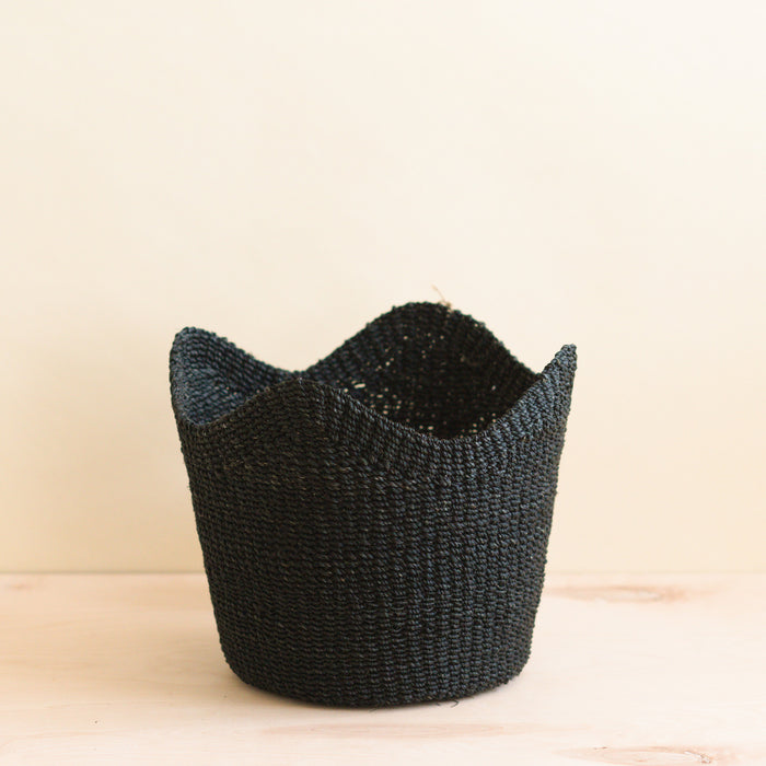 The LIKHA Abaca Scallop Basket, crafted from Manila hemp, features a black woven design with a wavy rim and is displayed on a light wooden surface against a plain beige background.