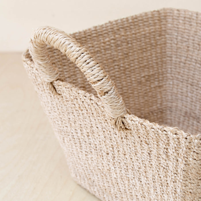 A close-up of the LIKHA Abaca Tapered Square Tabletop Basket with Handles, handwoven by Filipino artisans from natural abaca fiber, displayed on a light wooden surface.