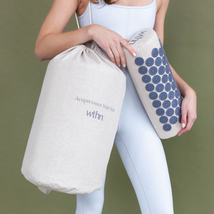 A person in a light blue bodysuit holds the Acupressure Mat Set by WTHN, perfect for relaxation and easing chronic pain, made from light beige fabric. One hand grips a cylindrical bag, while the other cradles a cushion with blue circular patterns. The WTHN logo contrasts against the olive green background.