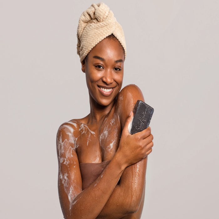 A smiling woman with a towel wrapped around her head holds a SheaMoisture African Black Soap Bar Soap with Shea Butter, washing her shoulder in rich lather from this natural skin cleanser against a plain background.