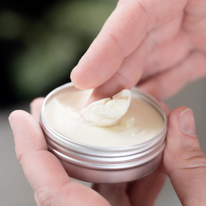 Close-up of someone using their fingers to scoop Lovett Sundries Aftershave Balm from a round tin. The blurred, greenish backdrop suggests a natural setting, fitting for this soothing balm to prevent razor burn.