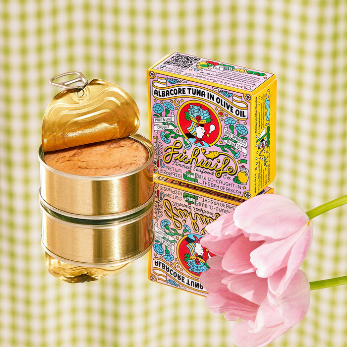 An open can of Fishwifes Albacore Tuna in Olive Oil showcases its vibrant contents, with vintage-style packaging evoking a trusted Spanish cannery. A pink tulip adorns the scene alongside a charming green and white checkered backdrop.