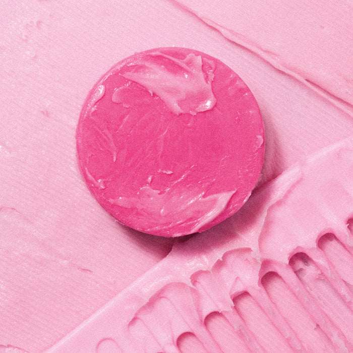 The ALINE Conditioner Bar from NOT!CE Hair Co is a round, creamy pink soap resting on a textured pink backdrop with brush strokes and a partially visible comb coated in the same luxurious pink conditioner.