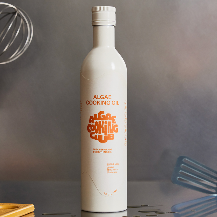 A white bottle labeled All Purpose Oil by Algae Cooking Club stands on a counter, featuring orange text and icons showcasing its high smoke point and Omega 9s, with kitchen utensils and oil drops visible in the background.