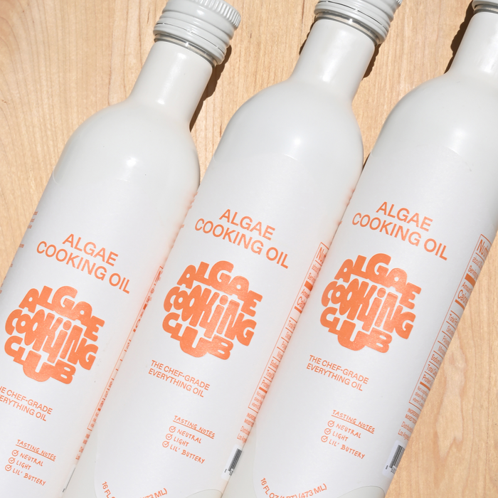 Three white bottles of Algae Cooking Club All Purpose Oil, a neutral oil with a 535°F smoke point, are arranged diagonally on a light wooden surface, showing their orange text and logos.