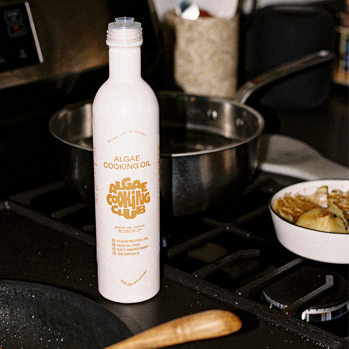 A bottle of Algae Cooking Club All Purpose Oil stands on a kitchen counter near a pan and cooked vegetables. The label highlights this clean, neutral oil as rich in Omega 9s, having a high smoke point, and being good for the future.