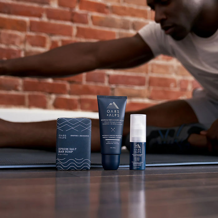 A man stretches on a yoga mat near a brick wall, with three Oars + Alps products—including Anti-Everything Body Powder—arranged in focus on the floor beside him.