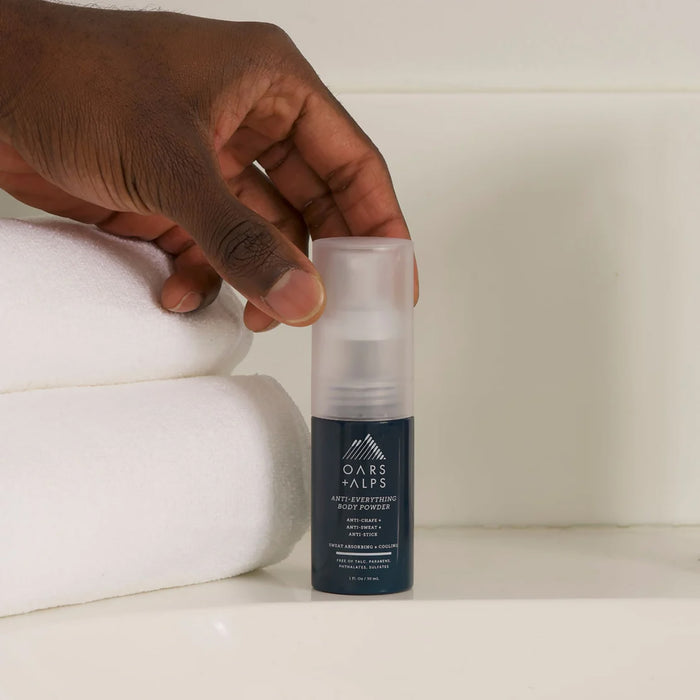A hand holding Oars + Alps Anti-Everything Body Powder, designed to absorb sweat and reduce friction, rests next to neatly folded white towels on a white surface.