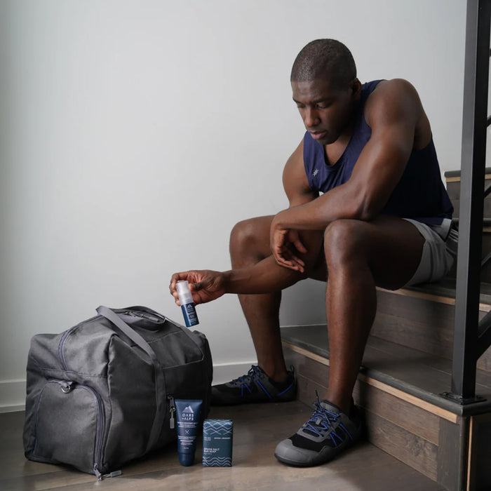 A man in athletic wear sits on wooden stairs, holding Oars + Alps Anti-Everything Body Powder, and looks at a gym bag. Other Oars + Alps grooming products, including one to absorb sweat and reduce friction, sit beside the bag.
