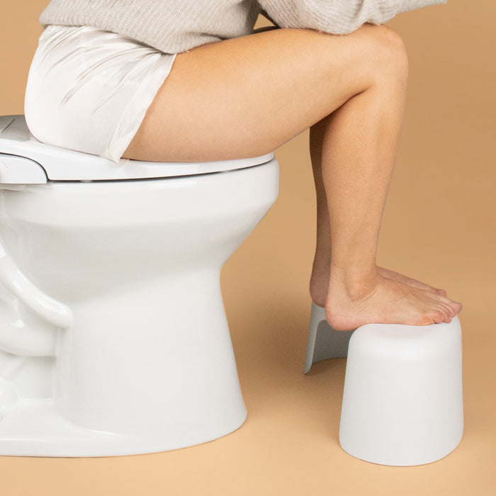 A person sits on a toilet using the TUSHY Arc Toilet Stool, demonstrating the squatting position for a simple bathroom upgrade, against a beige background.