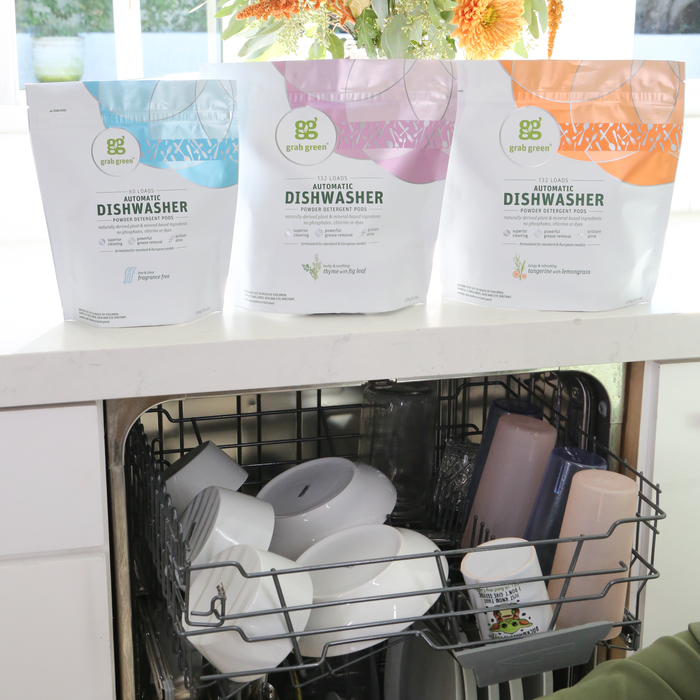 An open dishwasher displays sparkling white dishes and glassware. On the counter above, three grab green Automatic Dishwasher Detergent Pods bags in pink, green, and orange highlight their non-toxic formula. The window behind them shows a blurred view of greenery and orange flowers.