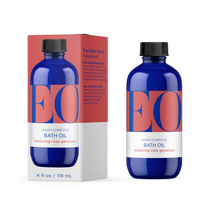 A 4 oz blue bottle of EO Products Bath Oil with calming chamomile rests next to its matching box, featuring a modern design and bold blue and pink text that emphasizes an aromatherapeutic bath ritual theme.