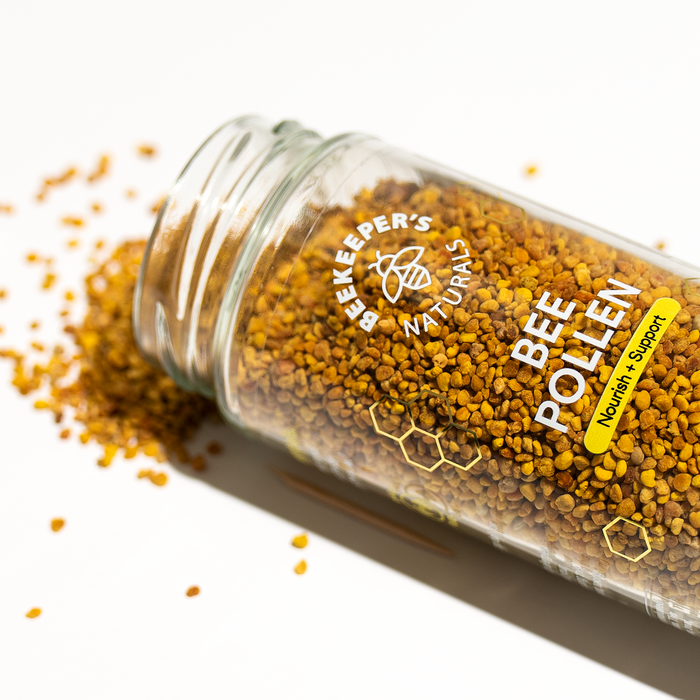 A tipped glass jar labeled Bee Pollen from Beekeepers Naturals spills golden granules, natures multivitamin, onto a white surface. The label says Nourish & Support and displays honeycomb graphics, highlighting its natural antioxidants.