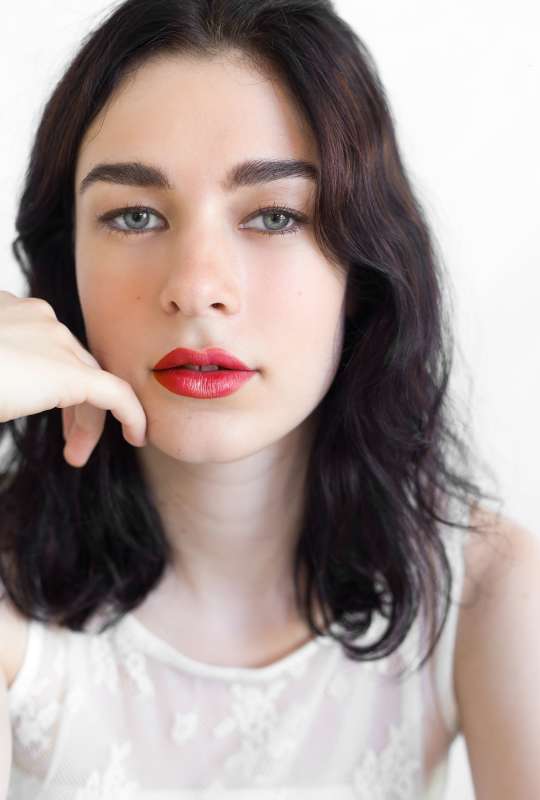 A person with shoulder-length dark hair and bright red Beetroot Cheek & Lip Tint by Ere Perez poses against a white background, resting their chin on their hand. They wear a sheer white lace top and have a neutral expression.