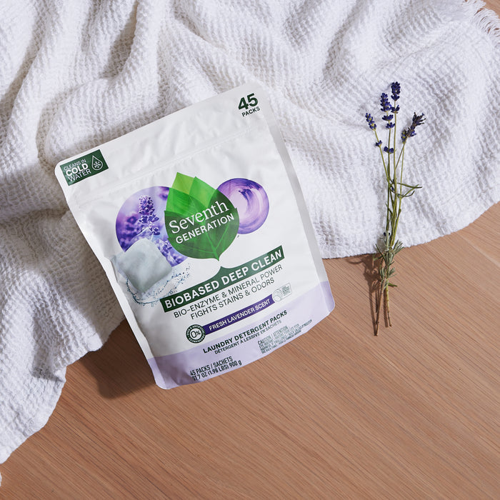 A bag of Seventh Generation Bio-Enzyme Laundry Detergent Packs, featuring the EPA Safer Choice mark, sits on a wooden surface. Its next to a white cloth and lavender sprig, with green and purple packaging holding 45 bio-enzyme packs.