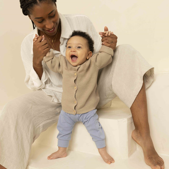 A smiling baby stands holding an adult’s hands. The adult sits barefoot on a step, both wearing neutral clothing. Nearby, ATTITUDE blooming belly Nursing Balm in zero-plastic packaging rests on the step.
