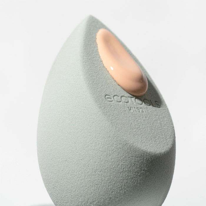Close-up of the EcoTools Blurring Blender, a light green, cruelty-free teardrop sponge ideal for a flawless finish. A drop of beige foundation rests on its textured surface, set against a soft white backdrop.