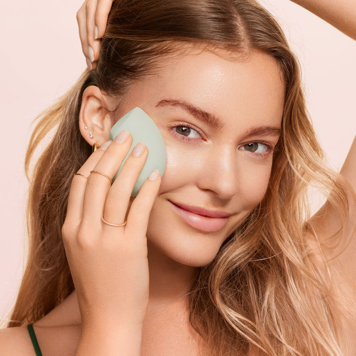 A woman with long blonde hair uses a green EcoTools Blurring Blender on her face, smiling gently. She wears delicate gold jewelry and a thin-strap top against a soft peach background, enhancing the serenity of this cruelty-free beauty ritual.