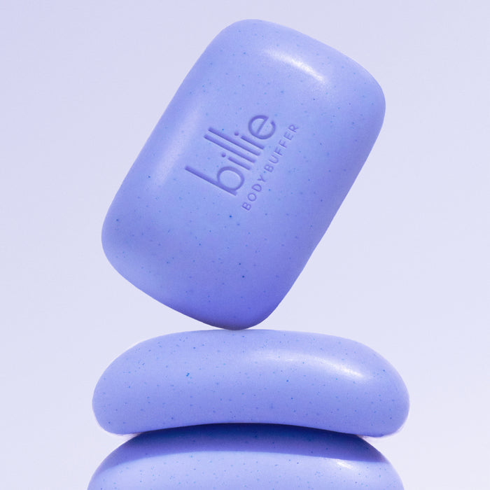 A purple bar of the billie Body Buffer - Pre-shave Exfoliating Bar, enriched with jojoba beads, balances on another identical bar against a light purple background.