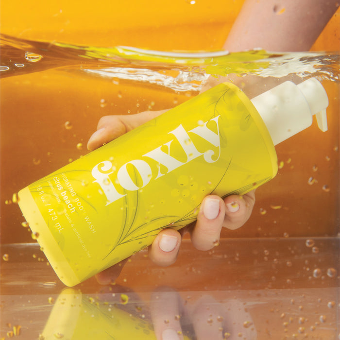 A hand partly submerged in warm yellow-orange water holds a Foxly Body Wash with pump dispenser and floral designs. The bottle stands out for its clean ingredients and elegant branding.