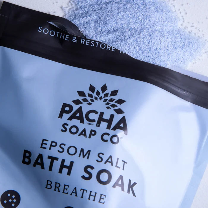 A partially open bag of Pacha Soap Co. Breathe Salt Soak rests on a surface, with light blue Epsom salt and the calming aroma of eucalyptus oil spilling out above the package.