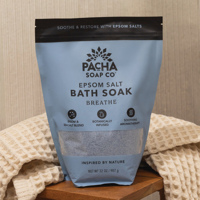 A bag of Pacha Soap Co. Breathe Salt Soak rests on a wooden surface beside a textured, cream-colored towel. Its blue and black packaging features crisp white text highlighting soothing Epsom salt and eucalyptus oil.