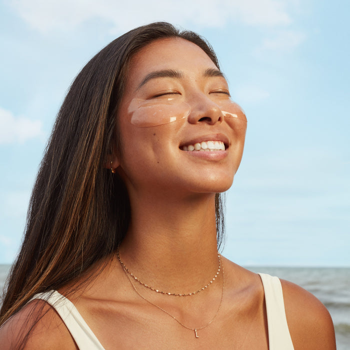A woman with long dark hair smiles with her eyes closed, wearing Three Ships Brighter Days Red Algae + Avocado Biodegradable Eye Masks outdoors by the ocean under a blue sky. She wears a light top and a delicate necklace.
