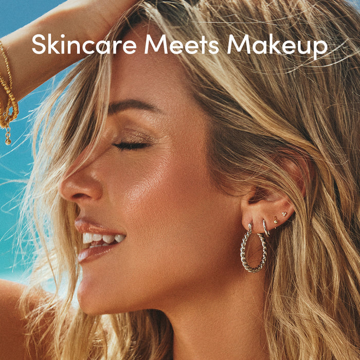 A woman with glowing, sun-kissed skin smiles with closed eyes, wearing hoop earrings. Text reads Uncommon Beauty Bronzing Drops: Skincare Meets Makeup on a sunlit beachy background for that post-vacation glow.