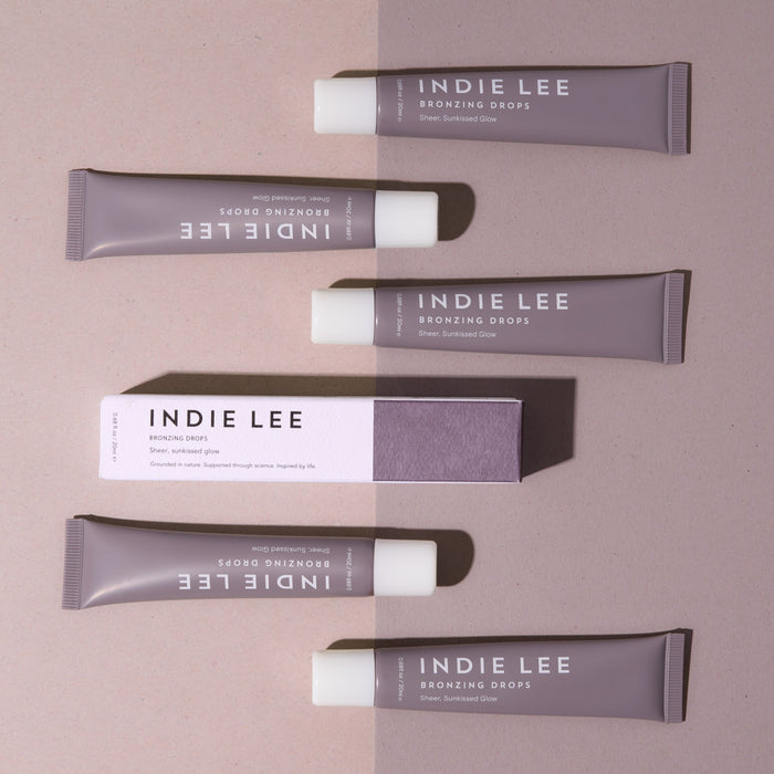 Five tubes and a box of Indie Lee Bronzing Drops are arranged neatly on a split beige and light brown background, with labels clearly visible—ideal for adding a hydrated summer glow.