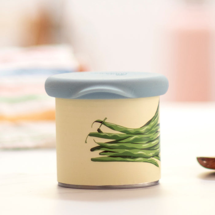 The Can Hugger by Food Huggers is a small, round reusable container featuring a light blue lid and cream-colored body with a green bean illustration. It rests on a white surface with blurred colorful objects behind and a wooden spoon to the right, perfect for eco-conscious pet owners.