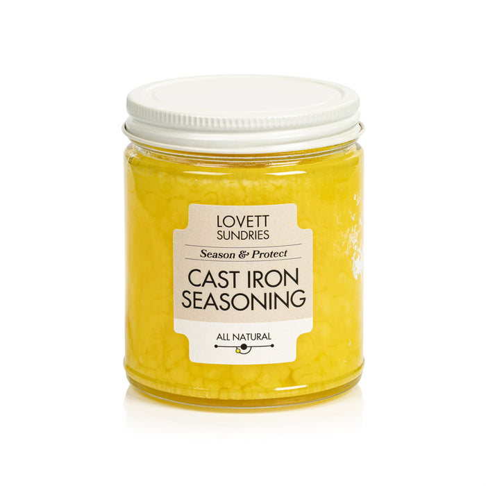 A clear glass jar with a white lid labeled Lovett Sundries Cast Iron Seasoning Oil. Inside is yellow, chunky all-natural oil to season and protect your cookware. The label reads Season & Protect and All Natural.