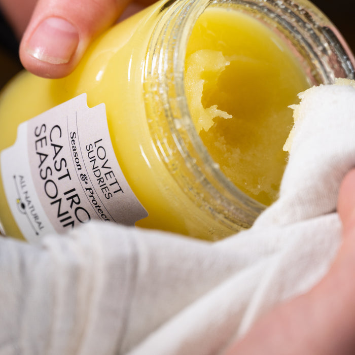 A hand scoops Lovett Sundries Cast Iron Seasoning Oil from a jar with a white cloth, ready to season cookware. The label hints at its all natural oils.