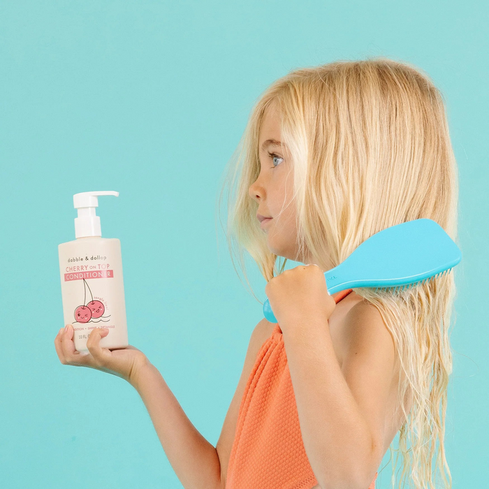 A young girl with long blonde hair stands against a turquoise background, wearing an orange top. She holds a bottle of Dabble & Dollops Cherry On Top Conditioner, known for deeply hydrating all hair types, and a blue brush in the other hand as she brushes her hair.