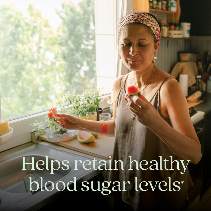 A woman stands in a sunlit kitchen, enjoying a piece of watermelon. Text on the image reads: Cinnamon Force Healthy Blood Sugar Level by New Chapter supports healthy blood sugar levels* with Non-GMO Cinnamon.