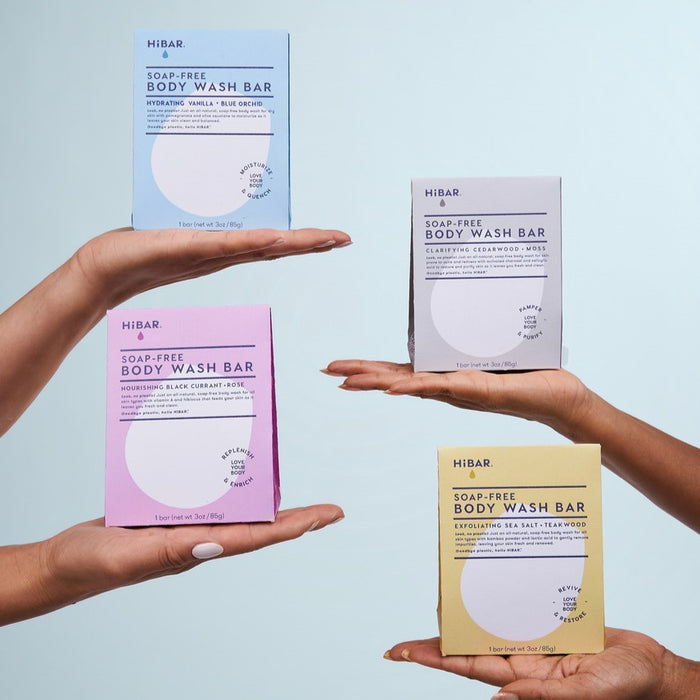 Four hands hold up HiBAR Clarify Body Wash Bars in colorful boxes against a light blue background. Each bar features a pH-balanced, non-comedogenic formula with unique scents and ingredients.