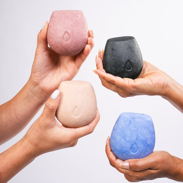Four hands each hold a rounded HiBAR Clarify Body Wash Bar in pink, black, beige, and blue against a white background. Each pH-balanced bar features a small droplet design on the front.