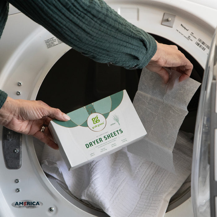 A person in a green sweater is putting a grab green Classic Dryer Sheet - Vetiver - NEW into a white front-loading washing machine filled with clothes to soften them and reduce static cling.