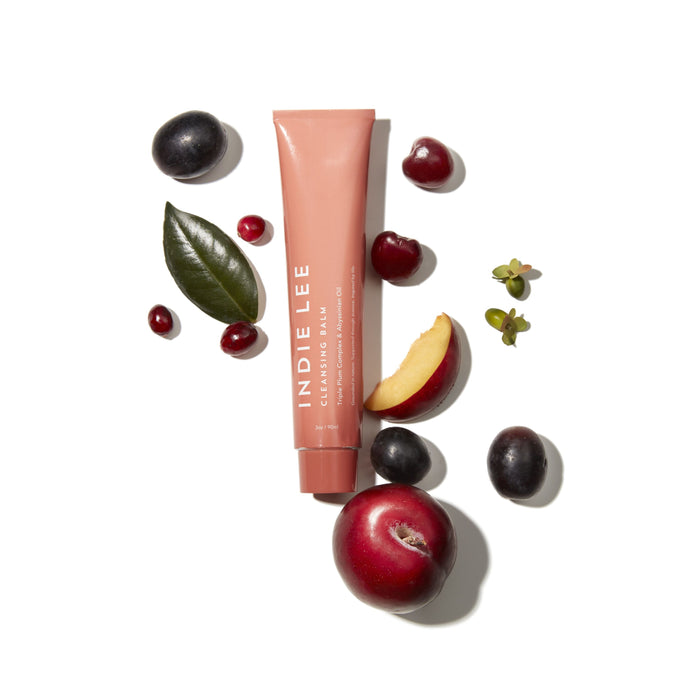Indie Lee Cleansing Balm, a vitamin C-rich balm, is artfully arranged on a white background with plums, plum slices, cherries, cranberries, a green leaf, and other small fruits.