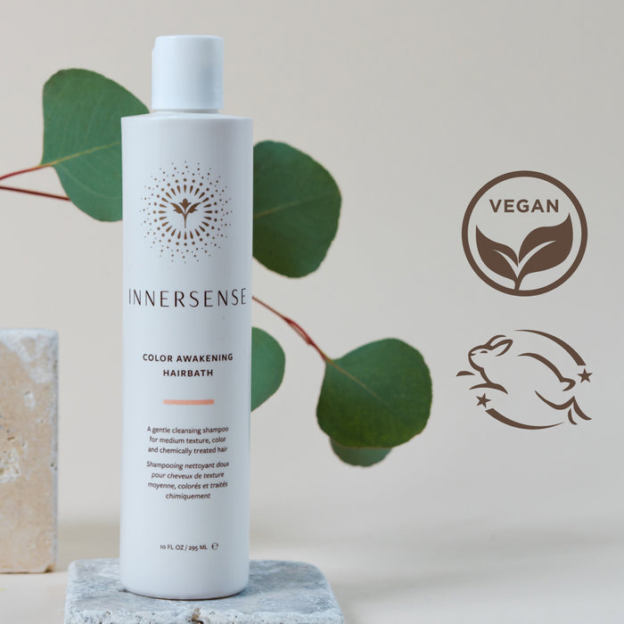 A bottle of Innersense Color Awakening Hairbath shampoo stands on a marble surface. Green leaves serve as the background. Two symbols nearby indicate the product is vegan and cruelty-free.